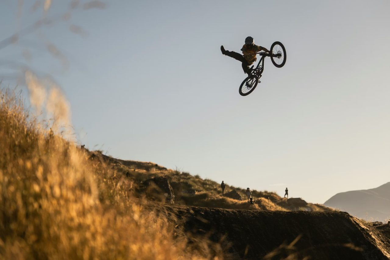 A mountain biker performing a mid-air trick over a dirt ramp during sunset, with a golden landscape in the background.