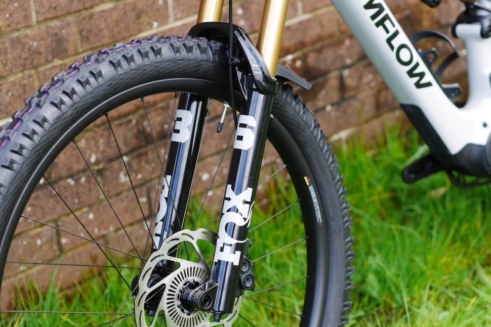 Close-up of a mountain bike front fork with FOX branding, featuring a black and gold design, disc brake, and rugged tyre against a brick wall background.