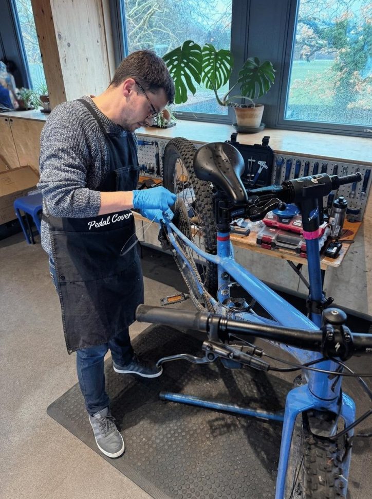 A mechanic wearing gloves and an apron is working on a blue bicycle in a workshop. The background features windows with natural light and indoor plants.