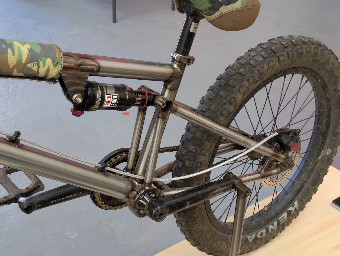 Close-up image of a bike's rear suspension and drivetrain, featuring a metallic frame, camouflaged seat, and a large, textured rear tyre.