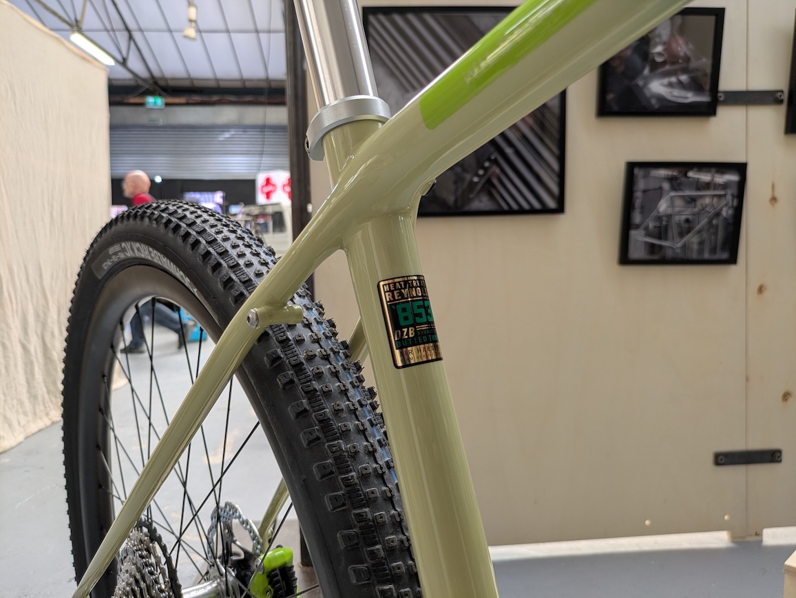 Close-up of a bicycle frame showing a digital display with a reading of 85%, alongside the tyre and suspension components, in an exhibition setting.