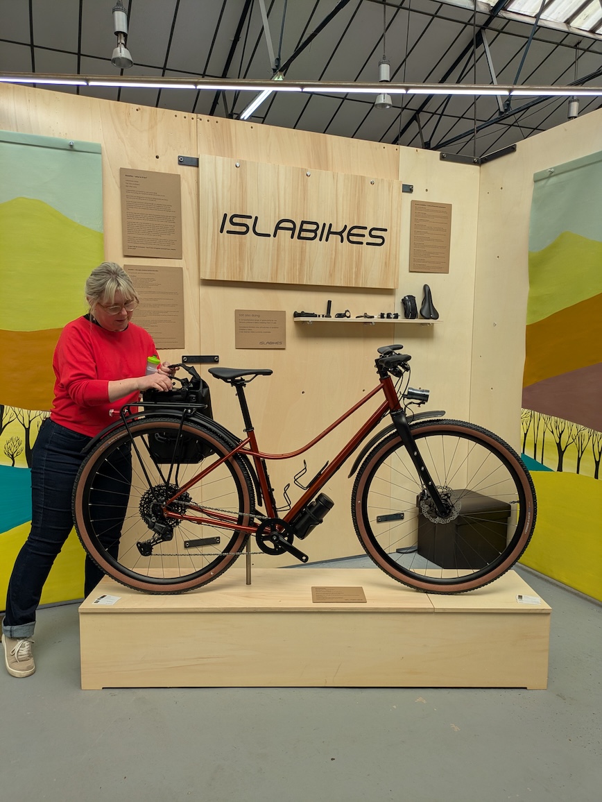 A woman in a red sweater is adjusting an urban bicycle displayed on a wooden stand, with an 'ISLABIKES' sign in the background and a colourful mural depicting hills.