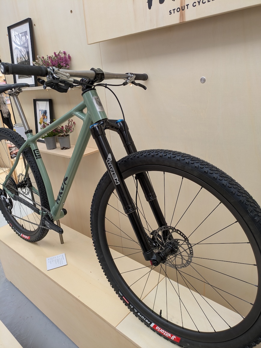 A green mountain bike displayed on a wooden pedestal, featuring a front suspension fork and knobby tyres, with a backdrop of framed pictures and potted plants.