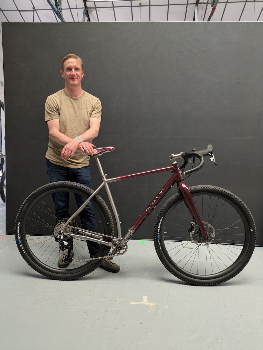 A man standing next to a metallic frame bicycle in a studio setting with a dark background.