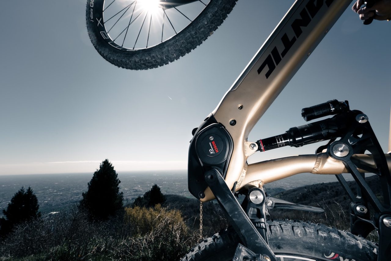 Close-up of a mountain bike's rear wheel and suspension system against a bright sky, showcasing the bike's components and elevated view of the landscape.