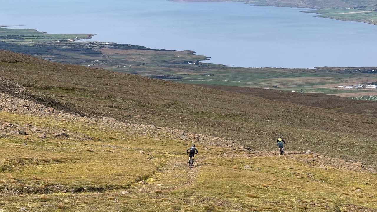 Two mountain bikers riding down a rocky trail with a view of a lake and farmland in the background.