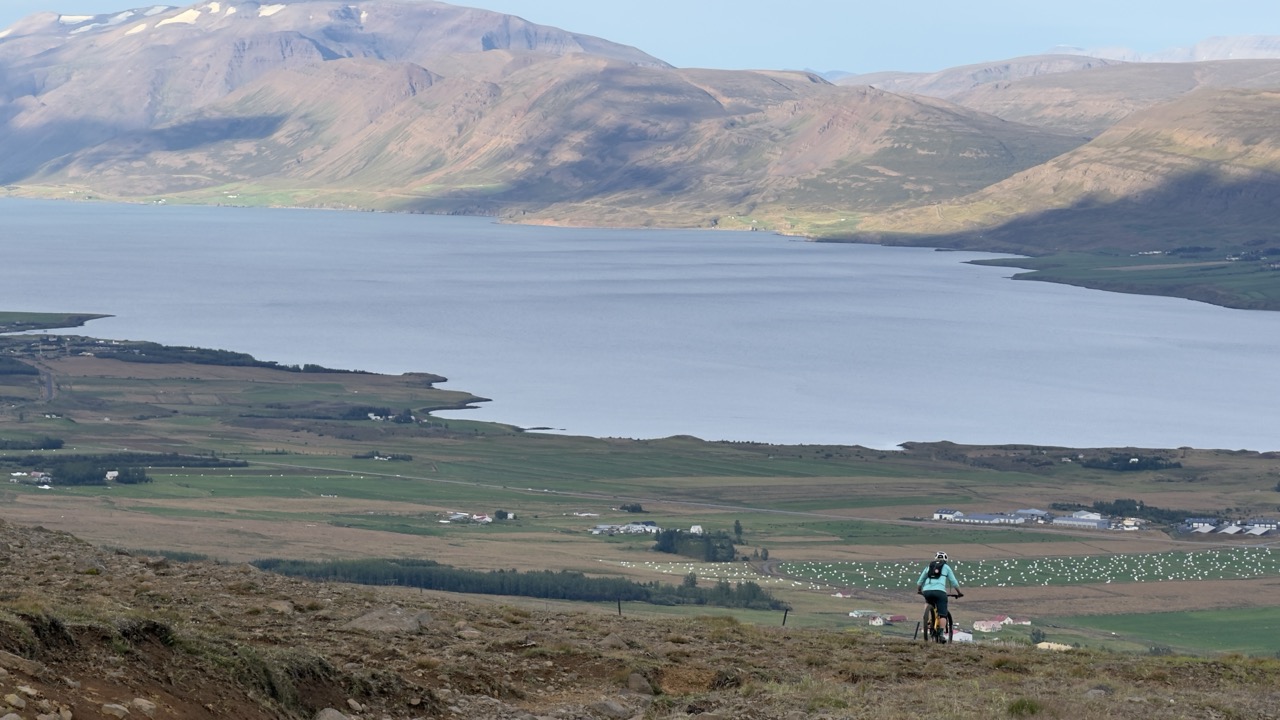 A cyclist riding on a mountain trail overlooking a scenic lake and surrounding countryside, with hills in the background.