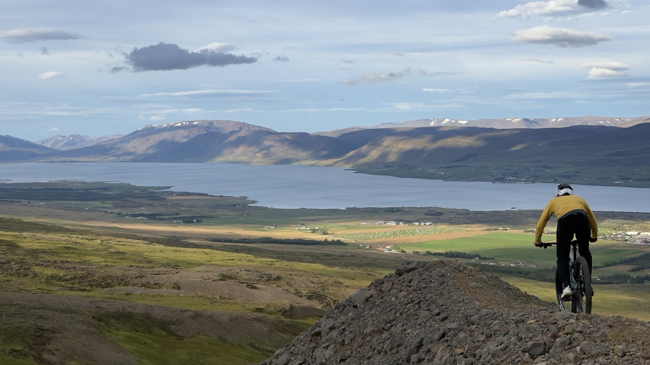 A mountain biker riding on a rocky trail overlooking a scenic landscape with a lake and mountains in the background. The sky is partly cloudy.