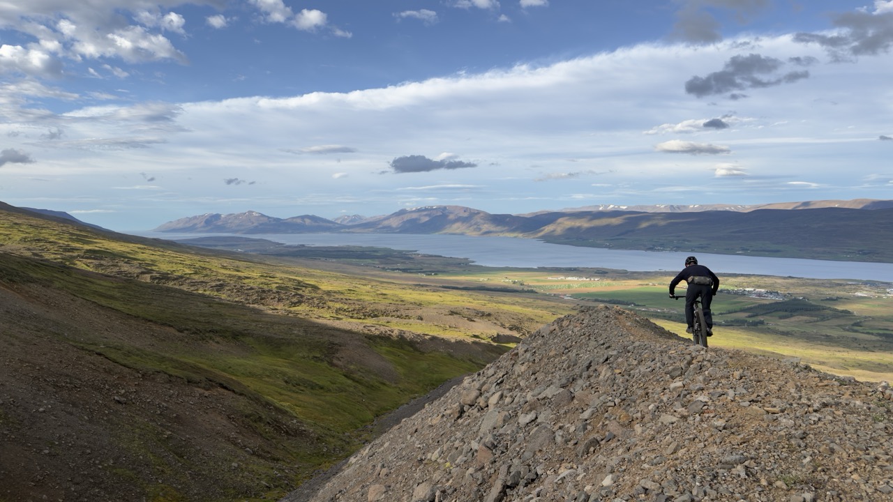 A mountain biker riding along a rocky ridge with a scenic view of valleys and mountains in the background under a partly cloudy sky.