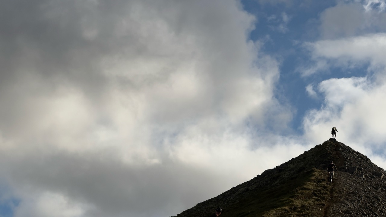 Two hikers ascending a steep, rocky hill under a cloudy sky.