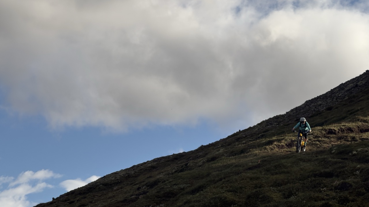 A mountain biker rides up a steep, grassy hill under a partly cloudy sky.