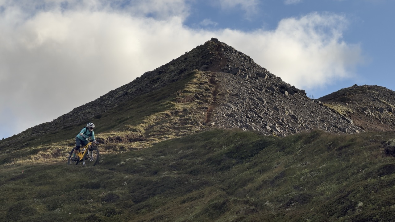 A cyclist riding a mountain bike down a grassy slope towards a rocky peak under a cloudy sky.