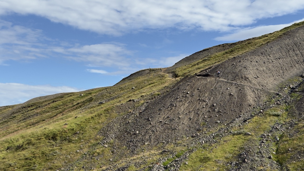 A cyclist navigating a rocky hillside with patches of grass under a partly cloudy sky.