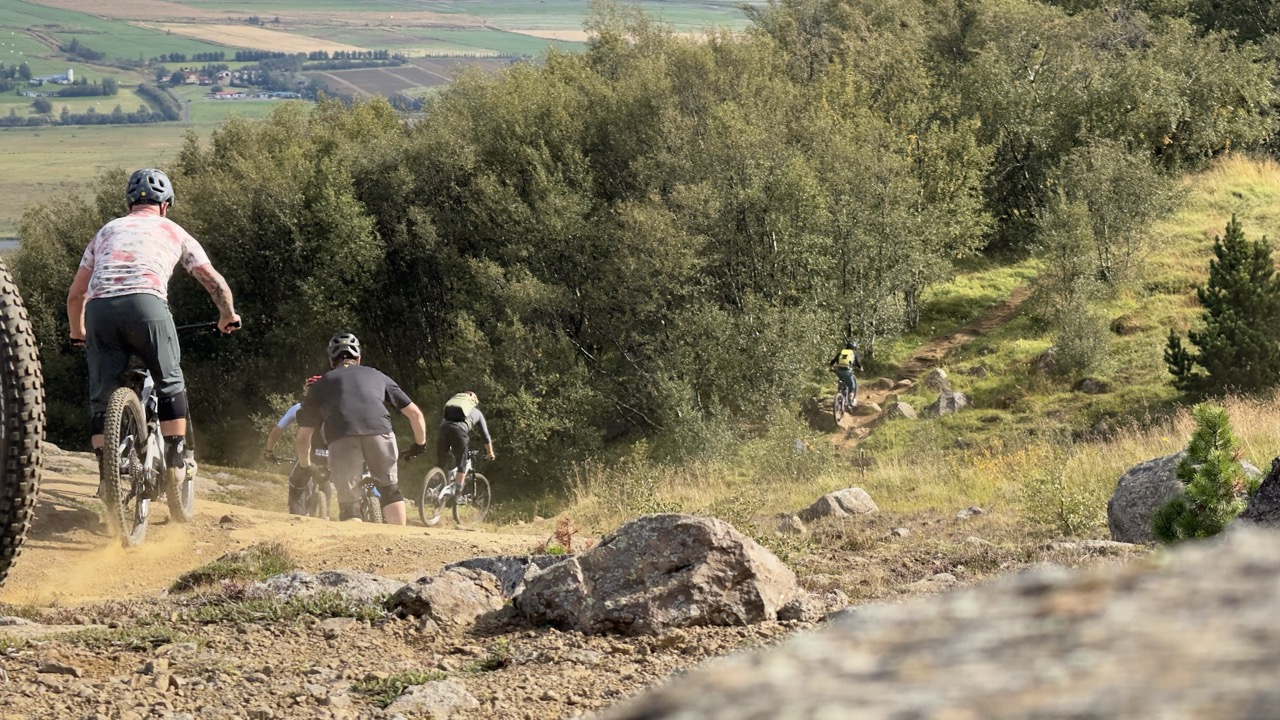 A group of mountain bikers riding on a dirt trail in a scenic landscape with greenery and fields in the background.