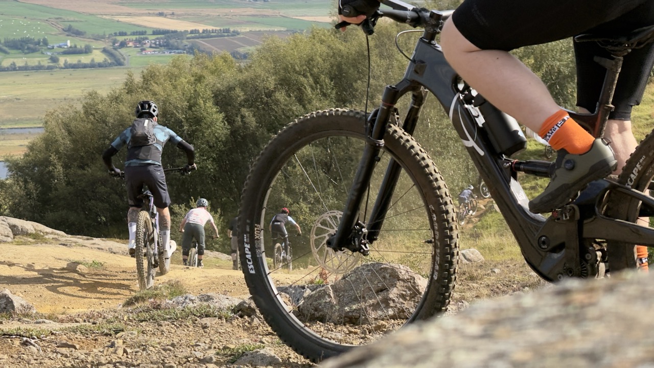 A group of mountain bikers riding on a dirt trail through a scenic landscape, with grassy hills and trees in the background.