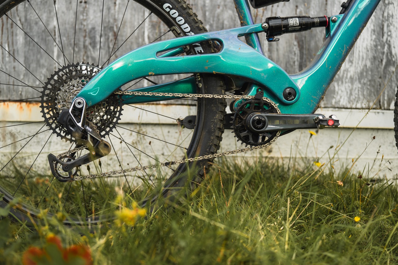Close-up of a mountain bike's drivetrain, featuring a turquoise frame, chain, and gears, set against a grassy background.