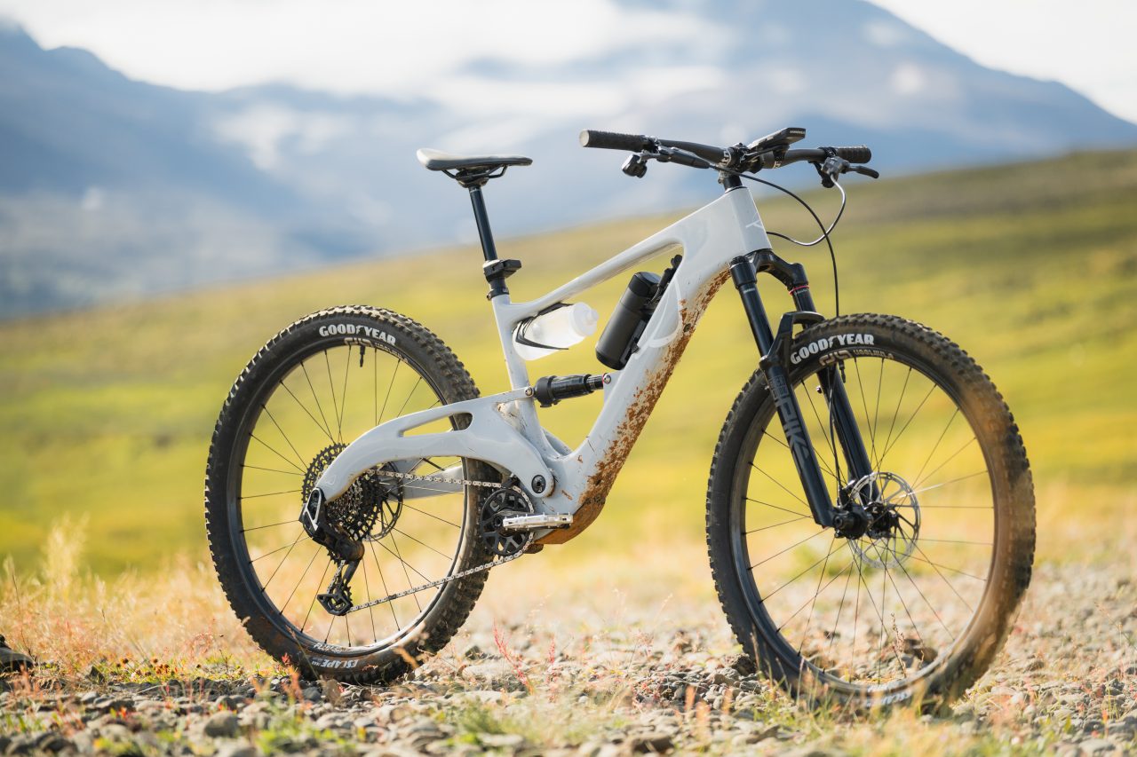 A modern mountain bike with a white frame and rugged tyres, positioned on a rocky surface with a green landscape and mountains in the background.