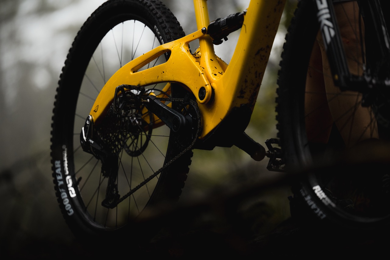 Close-up image of a yellow mountain bike frame and rear wheel in a forest setting, showcasing the chain and drivetrain components.