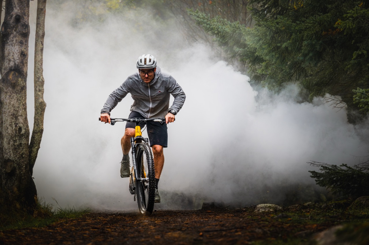 A cyclist in a grey jacket rides a mountain bike through a smoky forest trail, surrounded by trees.
